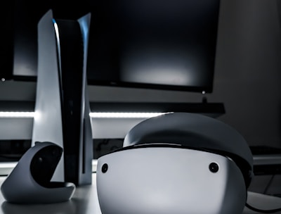 A sleek virtual reality headset sits on a reflective surface beside a gaming console and its controller. The background features a large dark monitor screen, creating a modern and futuristic setting. The lighting is subdued, highlighting the glossy surfaces of the electronic devices.